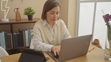 Orta yaşlı bir kadın iyi ışıklandırılmış, rahat bir ofiste dizüstü bilgisayarda dikkatle çalışır..