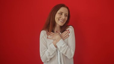 Beyaz bluzlu neşeli bir kızıl kadın canlı kırmızı bir arka plana karşı minnet dolu bir jest yapıyor..