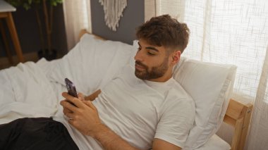 Yakışıklı İspanyol bir adam evinde rahat bir yatak odasında yatağında telefonuna bakıyor.