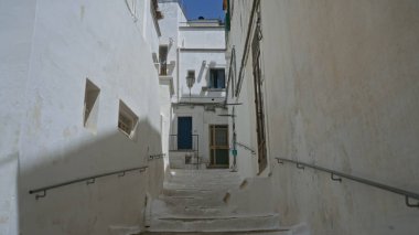 Dar, bembeyaz, ostuni, puglia, İtalya 'da basamakları olan dar bir sokak, berrak mavi gökyüzü altındaki geleneksel evlere açılıyor, Akdeniz mimarisi ve açık şehir cazibesi sergileniyor..