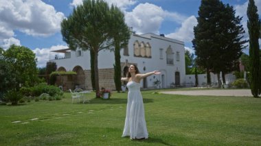 Genç bir İspanyol kadın, açık havada Puglia 'da güzel bir İtalyan villasının keyfini çıkarıyor. Parlak, bulutlu bir gökyüzünün altında selvi ağaçları olan büyüleyici beyaz bir evin önündeki yemyeşil çimlerin üzerinde duruyor..