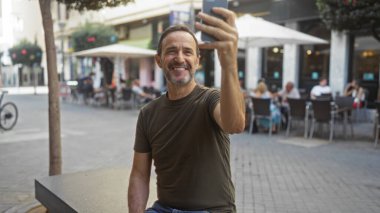 İspanyol bir adam kenar mahalle ortamında selfie çekiyor. Arka plandaki kafede yemek yerken mutlu bir şekilde gülümsüyor..