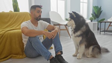 Yakışıklı genç İspanyol adam, köpeğiyle modern oturma odasında bağ kuruyor.