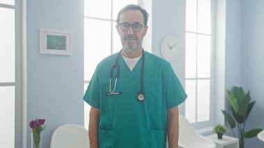 Önlüklü, olgun İspanyol erkek doktor güvenli bir hastane odasında duruyor..