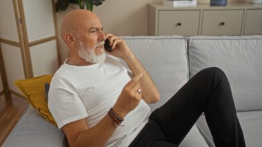 Oturma odasında kart tutarken telefon kullanan sakallı ve gri saçlı olgun bir adam. Kanepeye oturmuş, modern yaşam tarzını ön plana çıkarıyor.