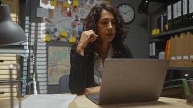 Focused woman detective working late on laptop in cluttered police station office with evidence board clipart