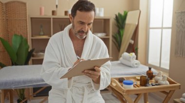 Sağlık merkezindeki olgun bir adam bornoz giyer ve panoya yazardı. Kapalı bir ortamda bitki ve güzellik ürünleri koyardı.