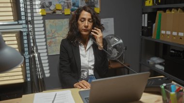 Takım elbiseli olgun bir kadın dedektif dağınık bir ofiste, elinde bir laptop ve kanıtlarla konuşuyor.