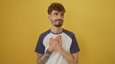 Sakallı ve dövmeli yakışıklı genç adam sarı arka planda düşünceli bir şekilde poz veriyor.