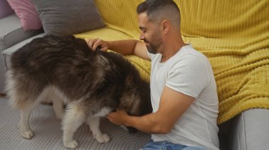 Neşeli, genç, İspanyol bir adam köpeğini rahat bir oturma odasında besliyor..