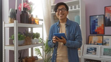 Elinde tablet tutan genç adam vazolar, bitkiler ve çerçeveli resimlerle çevrili arka planda raflarla ev dekorasyonu dükkanında gülümsüyor..