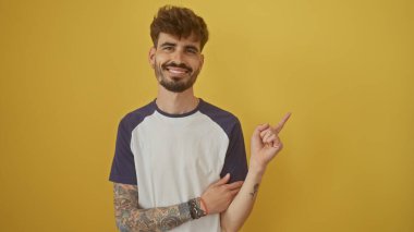 Yakışıklı, sakallı, İspanyol bir adam sarı bir arka planı işaret ediyor ve gülümsüyor..