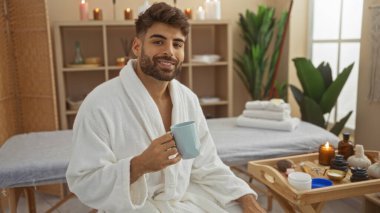 Yakışıklı bir adam huzurlu bir sağlık merkezinde spa tedavisi görüyor, beyaz bir bornoz giyiyor, elinde bir fincan çay tutuyor, mumlarla çevrili, ve ahşap tepside gerekli yağları taşıyor.