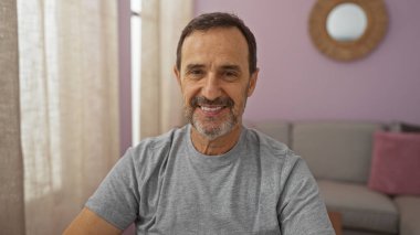 Orta yaşlı İspanyol bir adam, rahat bir oturma odasında gülümsüyor. Tarafsız dekor ve güneş ışığı perdelerin arasından akıyor..