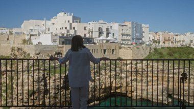Genç bir İspanyol kadın, güzel güneşli bir günde İtalya 'nın Puglia şehrinde, polignano' nun manzaralı uçurumlarına ve sahil binalarına bakan bir bakış açısında duruyor..