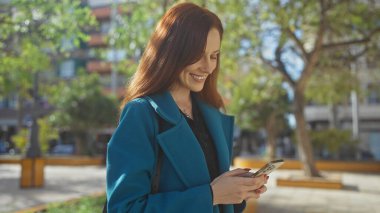 Deniz mavisi ceketli gülümseyen genç bir kadın güneşli bir şehir parkında akıllı telefon kullanıyor..