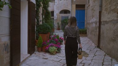 Çizgili gömlekli genç bir kadın polignano 'da büyüleyici, dar bir sokakta yürüyor. Bir kısrak, İtalya, canlı çiçekler ve kırsal mimariyle süslenmiş..