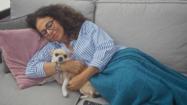 Olgun bir kadın, küçük chihuahua 'sıyla kapalı bir kanepede sarılır, sıcak, samimi, evcil bir sahneyi somutlaştırır..