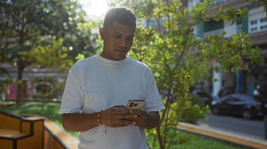 Beyaz tişörtlü genç İspanyol adam şehir parkında arka planda ağaçlar ve binalarla akıllı telefon kullanıyor.