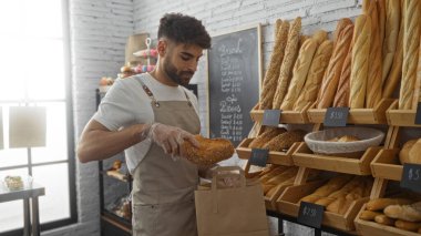 Fırında sakallı genç bir adam, ekmeği kese kağıdına yerleştirirken modern kapalı dükkanda çeşitli somunlar ve fiyat etiketli pastalarla çevrili duruyor.