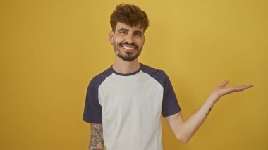 Smiling young man with beard and tattoos extends his hand against a yellow wall background in a casual pose.