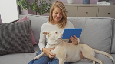 Kapalı bir koltukta köpeğiyle oturan gülümseyen bir kadın, sıcak bir dairede tablet tutuyor..