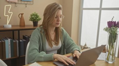 Kitaplar ve dekorlarla dolu rahat bir ofiste dizüstü bilgisayarla çalışan odaklanmış genç bir kadın..