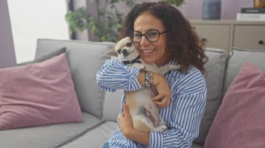 Gülümseyen orta yaşlı bir kadın sıcak bir oturma odasında bir chihuahua tutuyor..