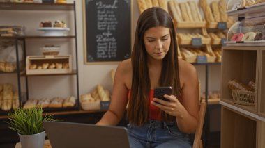 İspanyol kadın fırında akıllı telefon kullanırken arka planda ekmek ve hamur işleriyle dolu raflarda oturuyor..