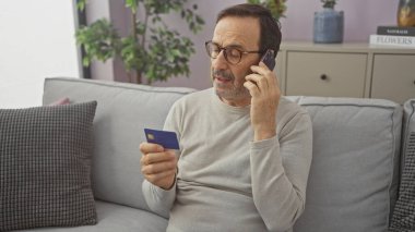 Akıllı telefondan konuşan ve oturma odasında kredi kartı tutan olgun bir İspanyol.
