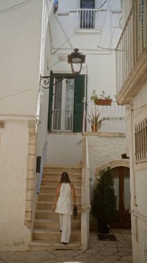 Beyaz elbiseli genç bir İspanyol kadın locorotondo, puglia, İtalya 'nın büyüleyici eski sokaklarında taş basamaklardan yukarı doğru yürür. Beyaz taş binalar ve yemyeşil bitkilerle çevrilidir..