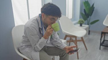 A young man in a white coat, possibly a healthcare professional, sipping from a mug and looking at a smartphone in a bright, plant-decorated room. clipart