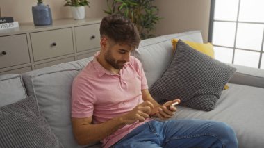 Oturma odasındaki koltukta oturan sakallı yakışıklı İspanyol bir adam akıllı telefon kullanıyor. Arka planda bitkiler ve dekorlar var. Pembe gömlek ve kot pantolon giymiş..