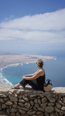 Lanzarote 'u gezen genç bir kadın, kanarya adaları, taştan bir duvarda oturuyor kıyı şeridi ve okyanus manzaralı, güneşli bir açık hava gününün tadını çıkarıyor..