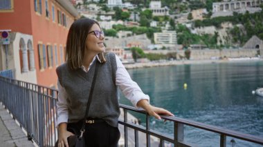 İspanyol kadın Villlefranche, Fransa 'da manzaralı Avrupa kıyı manzarasını seyrediyor, arka planda büyüleyici binalarla deniz kenarında gülümsüyor, genç seyahat zarafeti sergiliyor..