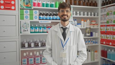 Beyaz önlüklü, gülümseyen sakallı bir eczacı tıbbi malzemeler ve hijyen ürünleriyle dolu bir eczanede güvenle duruyor..