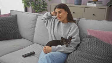 Genç bir kadın sıcak bir oturma odasındaki kanepede Chihuahua 'sıyla rahatlıyor, ev ve rahatlık hissi uyandırıyor..