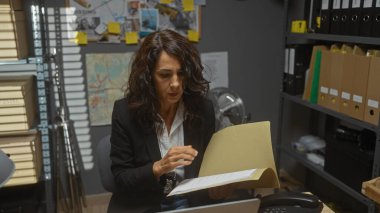 Olgun bir kadın dağınık bir dedektiflik bürosunda çalışkan bir şekilde evrakları inceliyor..