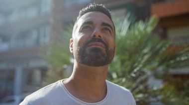 Düşünceli bir İspanyol 'un portresi yukarı bakıyor, güneşli bir şehir ortamında yeşil yeşillikler ve bulanık binalarla..