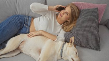 Genç bir kadın evdeki kanepede uzanıyor, rahat bir dairede telefonla konuşurken bir köpeği okşuyor..