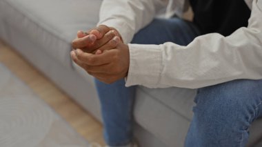Genç, Afro-Amerikan bir adam, günlük giysiler içinde, bir kanepede oturuyor, ellerini kenetliyor.