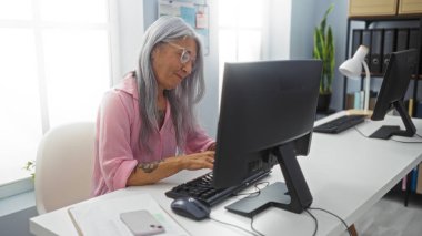 Geniş pencereli, parlak, modern bir ofiste bilgisayar başında çalışan, gülümseyen ve işine odaklanmış yaşlı bir kadın.