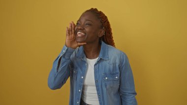Kot ceketli gülümseyen Afro-Amerikalı bir kadın sanki canlı sarı bir arka plana bağırıyormuş gibi el kol hareketi yapıyor.