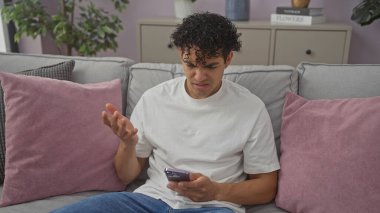 Kafası karışmış genç bir İspanyol adam, kanepede oturuyor ve modern bir dairede akıllı telefonuna bakıyor..