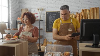 Bir pastanede birlikte çalışan erkek ve kadın fırında yemek sergiliyorlar ve arka planda ekmek ve hamur işleriyle birlikte kapalı bir tablette siparişleri kontrol ediyorlar.