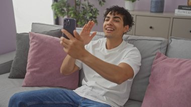Genç İspanyol bir adam, samimi bir oturma odasından, dostça bir el sallayarak neşeyi ifade eden bir video görüşmesi yapıyor..