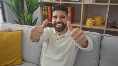 Sakallı, neşeli, genç, İspanyol bir adam sıcak, modern bir oturma odasında baş parmağını kaldırıyor..