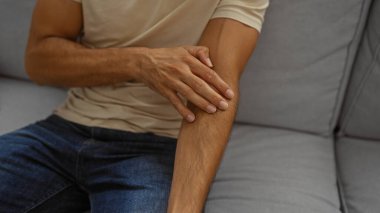 Genç bir adam, rahat bir oturma odasında kolunu kaşıyarak, günlük giysiler giyerek, iç mekandaki bir deri rahatsızlığını vurguluyor..