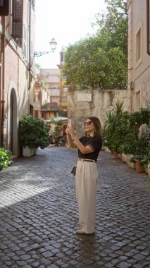 Roma, İtalya 'da bir sokakta, güneşli bir gökyüzünün altında, tarihi binalar ve kaldırım taşlarıyla çevrili eski kasaba cazibesini akıllı telefonuyla yakalayan bir kadın..