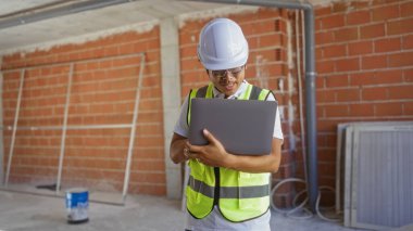 İnşaat alanında elinde dizüstü bilgisayarla oturan genç bir adam. Kasket ve güvenlik yeleği giyiyor.